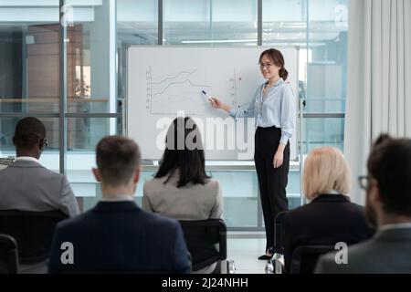 Junge, erfolgreiche Maklerin zeigt auf Finanzdiagramme auf dem Whiteboard, während sie vor dem Publikum im Hörsaal steht Stockfoto