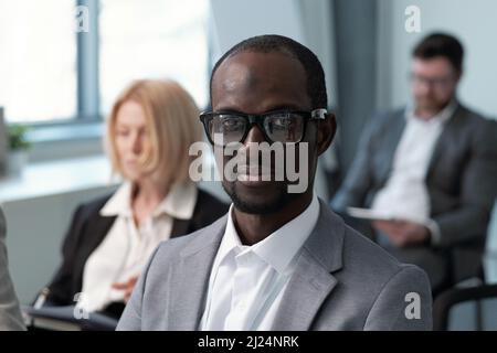 Junger zeitgenössischer schwarzer Mann in formalwear und Brillen, der auf die Kamera schaut, während er gegen männliche und weibliche Kollegen sitzt Stockfoto