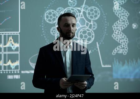 Ernsthafter männlicher Makler in formalwear, der Tablet hält, während er dem Publikum auf einer Konferenz oder einem Seminar über eine interaktive Tafel Bericht erstattet Stockfoto