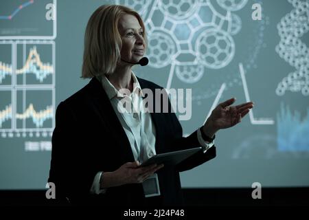 Glückliche blonde reife Geschäftsfrau mit Tablet Rede auf der Konferenz, während sie gegen interaktive Tafel stehen Stockfoto