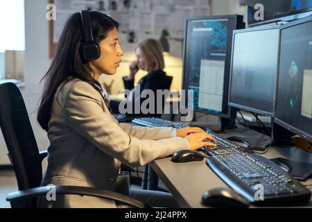 Seitenansicht einer jungen hispanischen Bedienerin des Überwachungssystems, die am Schreibtisch vor Computermonitoren im Büro sitzt Stockfoto