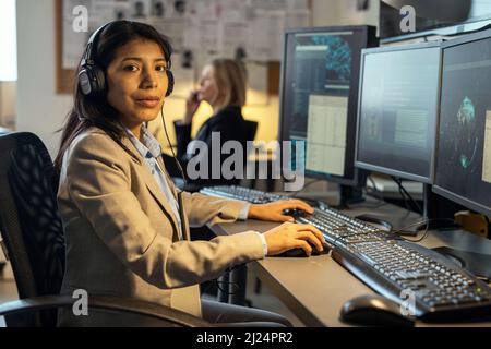 Junge, selbstbewusste hispanische Geheimagentin mit Kopfhörern, die während ihrer Arbeit im Büro auf die Kamera schauen Stockfoto