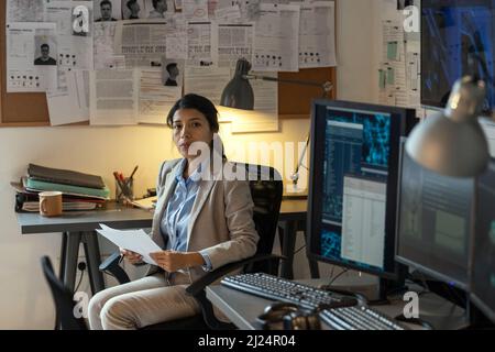 Junge, seriöse hispanische Angestellte der Sicherheitsabteilung mit Dokumenten, die auf die Kamera blicken, während sie in einem Sessel am Arbeitsplatz sitzen Stockfoto