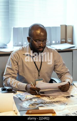 Ernsthafter männlicher FBI-Agent in formeller Kleidung, der sich darauf konzentriert, kriminelle Profile zu lesen, während er mit Dokumenten und Beweisen am Schreibtisch sitzt Stockfoto