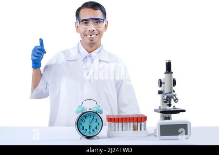 Asiatischer Nerd-Wissenschaftler, der mit einem Mikroskop und einem medizinischen Reagenzglasgestell auf dem Schreibtisch auf weißem Hintergrund isoliert steht und den Daumen zeigt Stockfoto