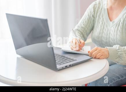 Seitenansicht und Nahaufnahme der Hände von Frauen mit Laptop-Bildschirm und Schreiben in Notizblock. Webinar. Praktische Arbeit mit digitaler Technologie Stockfoto