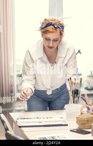 Eine junge, aktive Künstlerin mit roten Haaren, in einem weißen Hemd und Jeans mit einem Verband auf dem Kopf, malt am Tisch Stockfoto