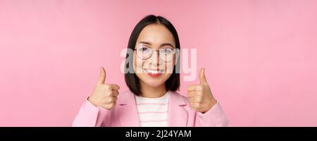 Super, Glückwunsch. Gesicht einer aufgeregten asiatischen Geschäftsfrau in einer Brille, lächelnd zufrieden, mit Daumen nach oben, stehend über rosa Hintergrund Stockfoto