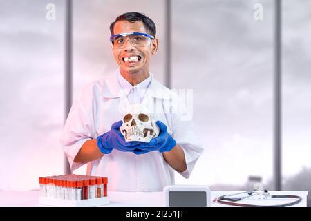 Asiatischer Nerd-Wissenschaftler, der im Labor einen Schädelkopf mit Stethoskop und medizinischem Röhrengestell auf dem Schreibtisch hält Stockfoto