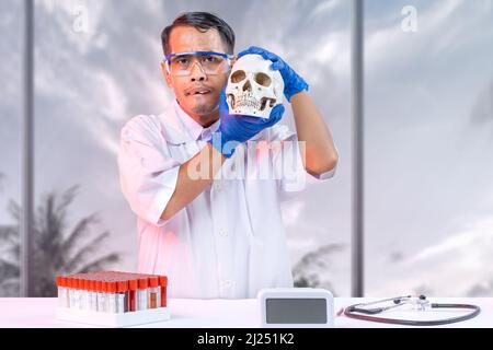 Asiatischer Nerd-Wissenschaftler, der im Labor einen Schädelkopf mit Stethoskop und medizinischem Röhrengestell auf dem Schreibtisch hält Stockfoto