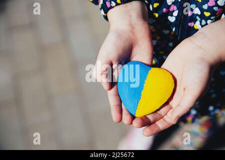 Kleines Kind halten herzförmigen Stein mit Ukraine blau-gelbe Nationalflagge oder Hymne. Mädchen lieben Heimat. Kinderstand für Ukrainer. Stoppt Krieg, Aggression, Invasion. Die Menschen verhals den Krieg. Stockfoto