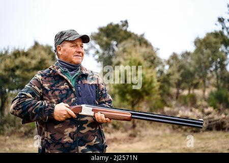 Männlicher Jäger in Camouflage-Oberbekleidung und Mütze, der eine Waffe trägt, während er im Herbstfeld herumhängt Stockfoto