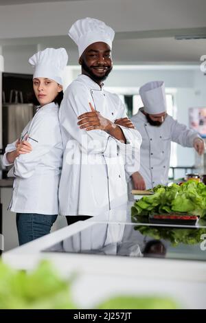 Multiethnische Gastronomieexperten stehen in der Restaurantküche mit gekreuzten Armen, während sie vor der Kamera posieren. Erfahrene Köche tragen Kochuniformen während der Zubereitung von frischem Gemüse für Gourmet-Rezept. Stockfoto