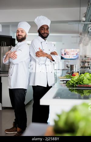 Porträt diverser kulinarischer Experten, die Kochhüte tragen, während sie in der Nähe der Kochstation stehen. Köche stehen in der professionellen Küche des Restaurants mit gekreuzten Armen, während sie Zutaten für das Gericht zubereiten. Stockfoto