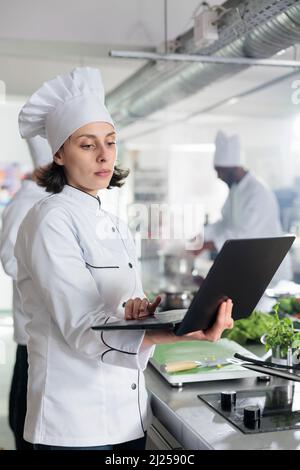 Restaurant Küchenarbeiter mit Laptop Suche Gourmet Küche Abendessen Gericht für Kochwettbewerb im Restaurant statt. Gastronomie-Experte mit Handheld-Computer auf der Suche nach Garnierung Ideen im Internet. Stockfoto