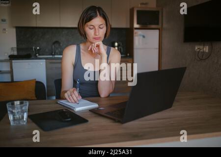 asiatische Freiberuflerin, die im Wohnzimmer an einem Laptop arbeitet. Fokussierte Dame, die entfernt studiert und Notizen im Copybook macht, Vortrag hört. Technologien für Online-Bildung und Selbstunterricht Stockfoto