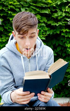 Überraschte den jungen Mann mit einem Buch auf dem Hintergrund der grünen Blätter Stockfoto