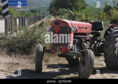 Ein alter roter klassischer Traktor links auf dem Boden Straße Seite in der Nähe einer Brücke mit einem Schild geschrieben Greenriver. Nostalgische Sicht auf die Landwirtschaft. Stockfoto