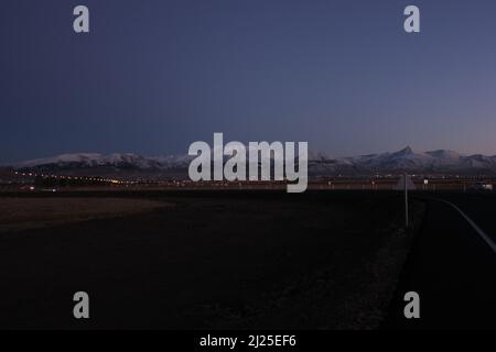 Kleine Stadt an den Hügeln von weißen schneebedeckten Bergen in der Morgendämmerung mit eingeschaltetem Straßen- und Hauslicht und dramatischer blau-grauer Himmelfarbe. Stockfoto