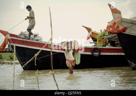 Ein Mann trägt sein Gepäck, während er auf flachem Wasser zwischen Booten läuft, die am Haldi-Fluss in Haldia, Purba Medinipur, Westbengalen, Indien ankerten. Laut einem von den Vereinten Nationen am 28. Februar 2022 veröffentlichten Bericht des Zwischenstaatlichen Ausschusses für Klimaänderungen werden in den nächsten 30 Jahren erstaunliche 143 Millionen Menschen durch steigende Meere, Dürre, sengende Temperaturen und andere Klimakatastrophen entwurzelt. Stockfoto