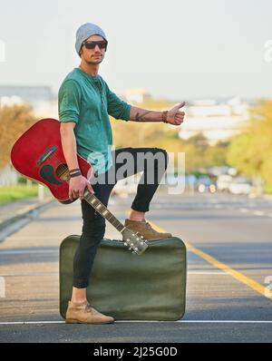 Mit seiner Musik unterwegs. Aufnahme eines hübschen jungen Mannes, der mit einer Gitarre und einem Koffer am Straßenrand trampt. Stockfoto