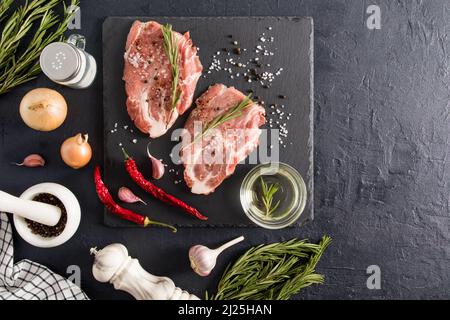 Draufsicht auf zwei saftige rohe Schweinesteaks mit Gewürzen auf einer Schiefertafel mit Zutaten zum Grillen. Schwarzer Hintergrund Stockfoto