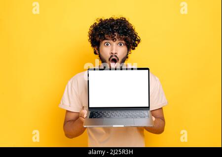 Schockiert erstaunt indischen oder arabischen lockigen Kerl, hält offenen Laptop, mit leeren weißen Bildschirm, Modell-Konzept, stand über isolierten orangefarbenen Hintergrund, Blick auf die Kamera mit offenem Mund überrascht Stockfoto