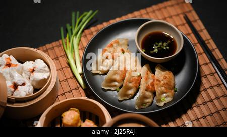 Gyoza auf einem schwarzen Teller mit spezieller Sauce, gedünstete chinesische Schweinebraten in einem Bambusdampfer auf Bambus-Tischset auf schwarzem Tisch. Asiatisch-chinesisches Speisekonzept Stockfoto