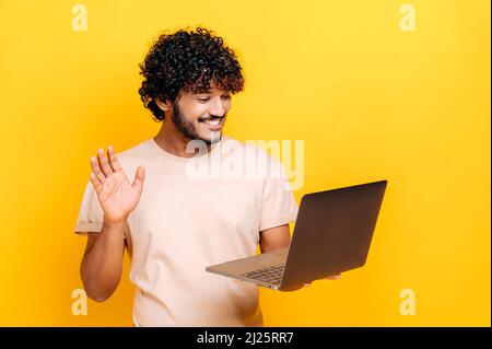 Positiver junger erwachsener inder oder araber, der Laptop hält, auf einer Online-Videokonferenz spricht, Gesprächspartner begrüßt, freundlich lächelt, auf einem isolierten orangefarbenen Hintergrund in einem lässigen T-Shirt steht Stockfoto
