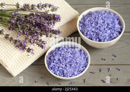 Meersalz mit Lavendel in einer weißen Schale und Lavendelblüten auf einem Leinentuch befinden sich auf einem grauen Holztisch. Nahaufnahme. Stockfoto