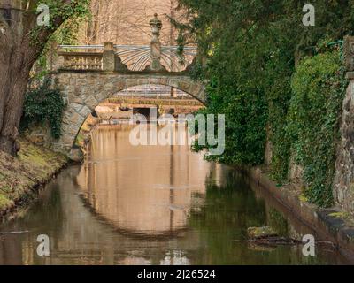 Ein kleiner Fluss, der durch den Park fließt. Es gibt eine gemauerte, historische Steinfußgängerbrücke über den Fluss. Es ist ein sonniger Tag. Stockfoto