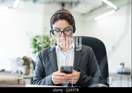 Der Callcenter-Betreiber langweilt sich und nutzt am Arbeitsplatz ein Smartphone. Eine Geschäftsfrau sitzt an ihrem Schreibtisch und tippt auf einem Mobiltelefon Stockfoto