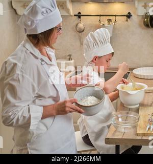 Mutter und Sohn kochen Apfelkuchen in der heimischen Küche. Eine Frau und ein Junge in Küchenhüten und Schürzen kochen mit Gebäck Stockfoto