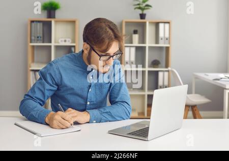 Konzentrierter junger Mann mit Brille macht sich Notizen, während er zu Hause mit dem Laptop arbeitet. Stockfoto