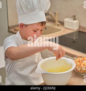 Ein Junge in der Kleidung des Küchenchefs gießt Äpfel in den Teig, um eine Torte zu kochen Stockfoto
