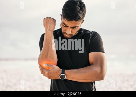 Ich denke, das ist mein Workout vorbei. Kurzer Schuss eines hübschen jungen Athleten, der seinen Ellbogen vor Schmerzen hält, während er am Strand trainiert. Stockfoto