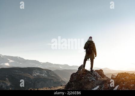 Die junge Wanderin steht auf einem großen Felsen vor der Kulisse der Berge und beobachtet den Sonnenuntergang Stockfoto