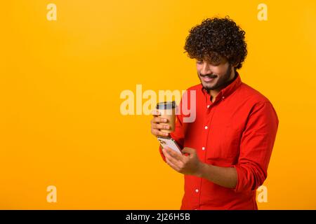Selbstbewusster Mann aus dem Nahen Osten trägt ein elegantes, legeres Hemd, das auf dem Smartphone SMS schreibt und eine Tasse Kaffee zum Mitnehmen hält. Männlicher Büroangestellter ist Stockfoto