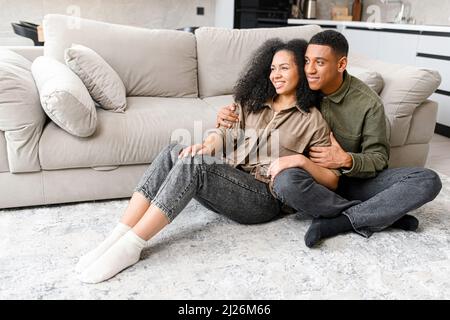 Lächelnder Mann umarmt sich hinter seiner charmanten Frau, während er sitzt und wegschaut. Junges multirassisches Paar, das zu Hause Zeit in einem gemütlichen, modernen Wohnzimmer verbringt Stockfoto