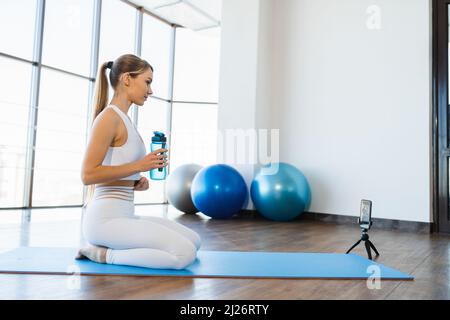 Junge Fitness-Frau sitzt auf einer Yogamatte mit einer Flasche Wasser und Live-Streaming-Workout Stockfoto