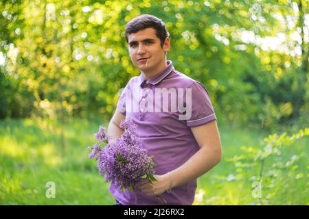 Defokussieren angenehm fröhlich schöner junger Kerl, der zur Seite schaut und einen Strauß Fliederblumen hält, während er sich glücklich fühlt. Junger schöner Mann mit Blumen. Stockfoto
