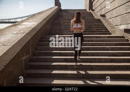 Sportliche Dame im modernen Trainingsanzug läuft Steintreppen am Stadtufer hinauf Stockfoto