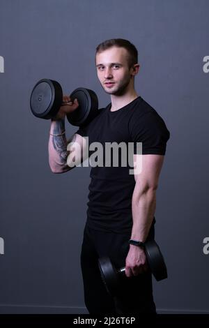 Ein Kerl mit Hanteln in einem schwarzen T-Shirt muskulösen Mann Hintergrund posiert, für mittlere Kopfhörer in stark für Sportler Gesundheit, Erwachsene t.. Shirt Bizeps Stockfoto