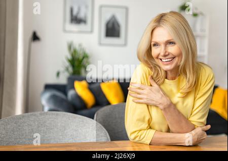 Nahaufnahme des Porträts einer blonden Frau in der Inneneinrichtung. Charmante reife kaukasische Dame, die wegschaut und lächelt, sich ausruhend, gelassene Frau, die das Wochenende zu Hause verbringt Stockfoto