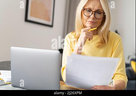 Konzentrierte ältere Geschäftsfrau macht Schreibarbeiten, während sie mit einem Laptop zu Hause am Tisch sitzt, fokussierte leitende Angestellte, die Dokumente vorbereitet und Rechnungen von zu Hause aus überprüft Stockfoto