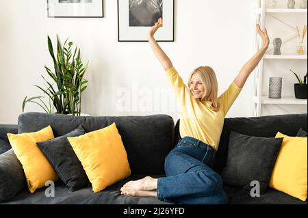 Eine unbeschwerte Frau mittleren Alters mit langen Haaren, die in einer modernen Wohnung auf dem Sofa sitzt, eine ältere Frau, die sich zu Hause ausruhte und eine träumende Anzeige wegschaute Stockfoto