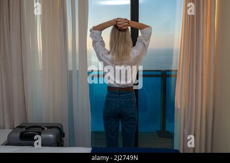 Eine junge Frau in einem weißen Hemd und einer blauen Jeans steht mit den Händen auf dem Kopf in einem Hotelzimmer am Fenster. Reisecncept und Sommerferien. Stockfoto