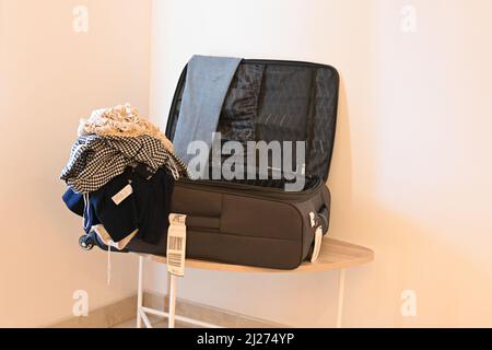 Ein offener Koffer in einer Hotelsuite, der auf einem Tisch sitzt, offen, mit Kleidung, die sich an der Seite stapelt. Stockfoto