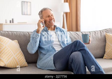 Fröhlicher, reifer Mann, der auf dem Sofa sitzt und mit dem Handy spricht Stockfoto
