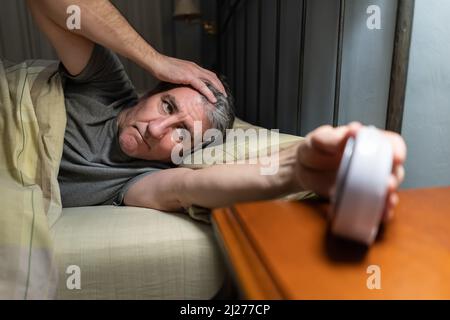 Mann mit Schlaflosigkeit liegt im Bett und schaut auf die Uhrzeit auf der Uhr. Stockfoto
